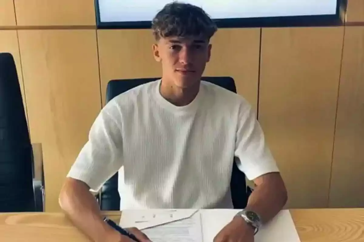 Joven sentado en una mesa firmando un documento con una pluma, vistiendo camiseta blanca y reloj plateado. Joven sentado en una mesa firmando un documento con una pluma, vistiendo camiseta blanca y reloj plateado.