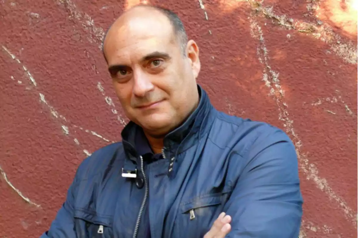 Bald man in a blue jacket posing with his arms crossed in front of a reddish wall Bald man in a blue jacket posing with his arms crossed in front of a reddish wall