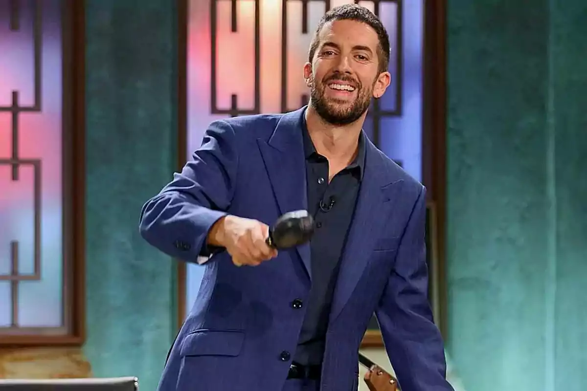 Hombre con barba y cabello corto usando un traje azul sonríe mientras sostiene un mazo en un set de televisión