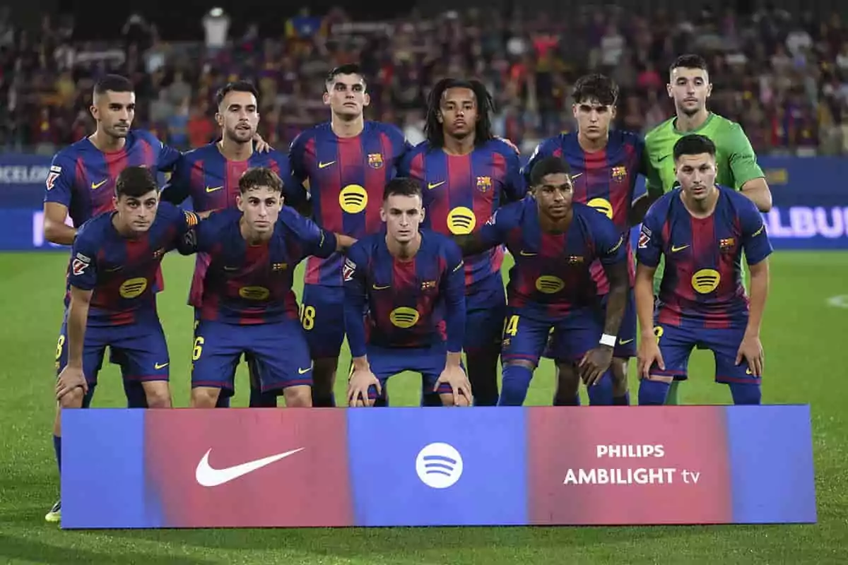 Once jugadores del FC Barcelona posan juntos en el campo antes de un partido con el uniforme azul y grana y una pancarta de patrocinadores frente a ellos Once jugadores del FC Barcelona posan juntos en el campo antes de un partido con el uniforme azul y grana y una pancarta de patrocinadores frente a ellos