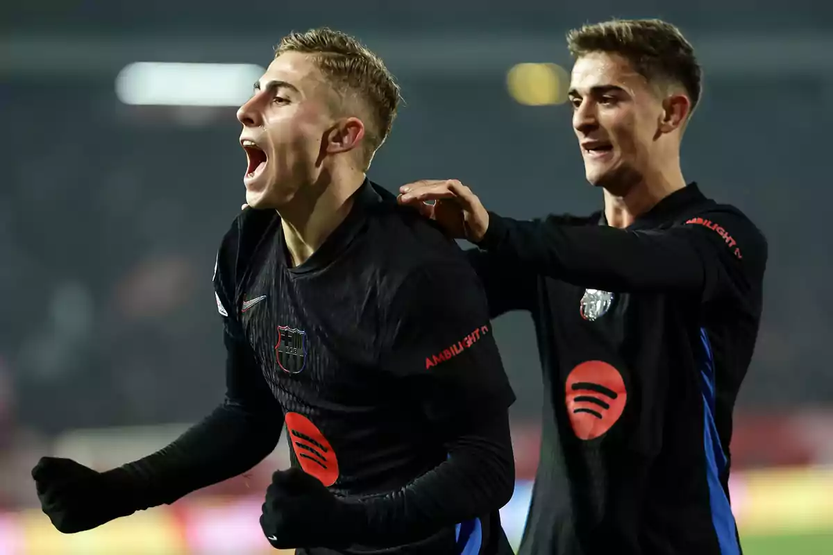 Two Barcelona players celebrate a goal enthusiastically during a match, both wearing black uniforms with red and blue details. Two Barcelona players celebrate a goal enthusiastically during a match, both wearing black uniforms with red and blue details.