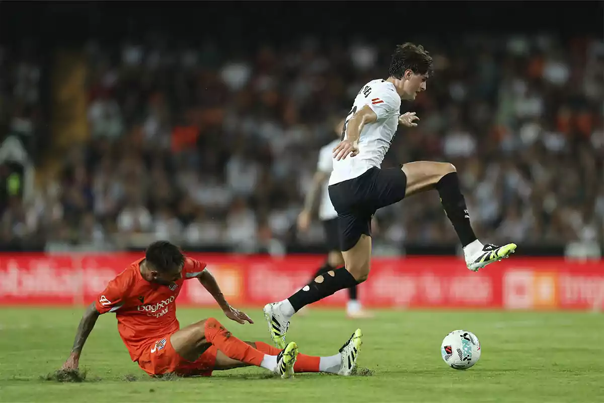 Un jugador de fútbol con uniforme blanco salta para evitar una barrida de un rival vestido de naranja durante un partido en un estadio lleno de espectadores.
