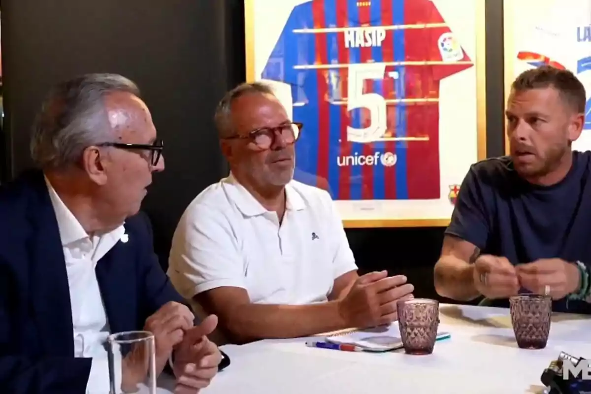 Tres hombres conversan sentados en una mesa con vasos y libretas, al fondo se observa una camiseta de fútbol enmarcada con el nombre Masip y el número 5.