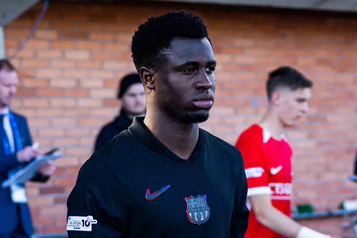 Jugador de fútbol con uniforme negro del FC Barcelona caminando en un estadio con fondo de pared de ladrillos y otras personas desenfocadas detrás Jugador de fútbol con uniforme negro del FC Barcelona caminando en un estadio con fondo de pared de ladrillos y otras personas desenfocadas detrás
