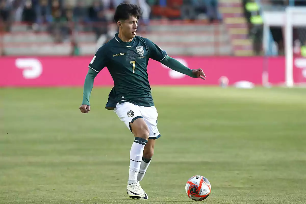 Futbolista con uniforme verde y blanco controlando un balón en un campo de fútbol durante un partido