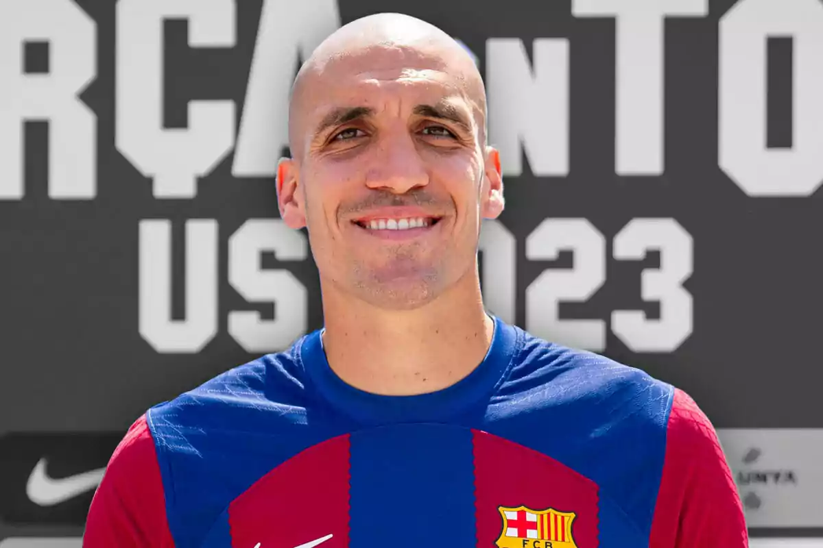 Bald man smiling wearing an FC Barcelona jersey in front of a background with large white letters