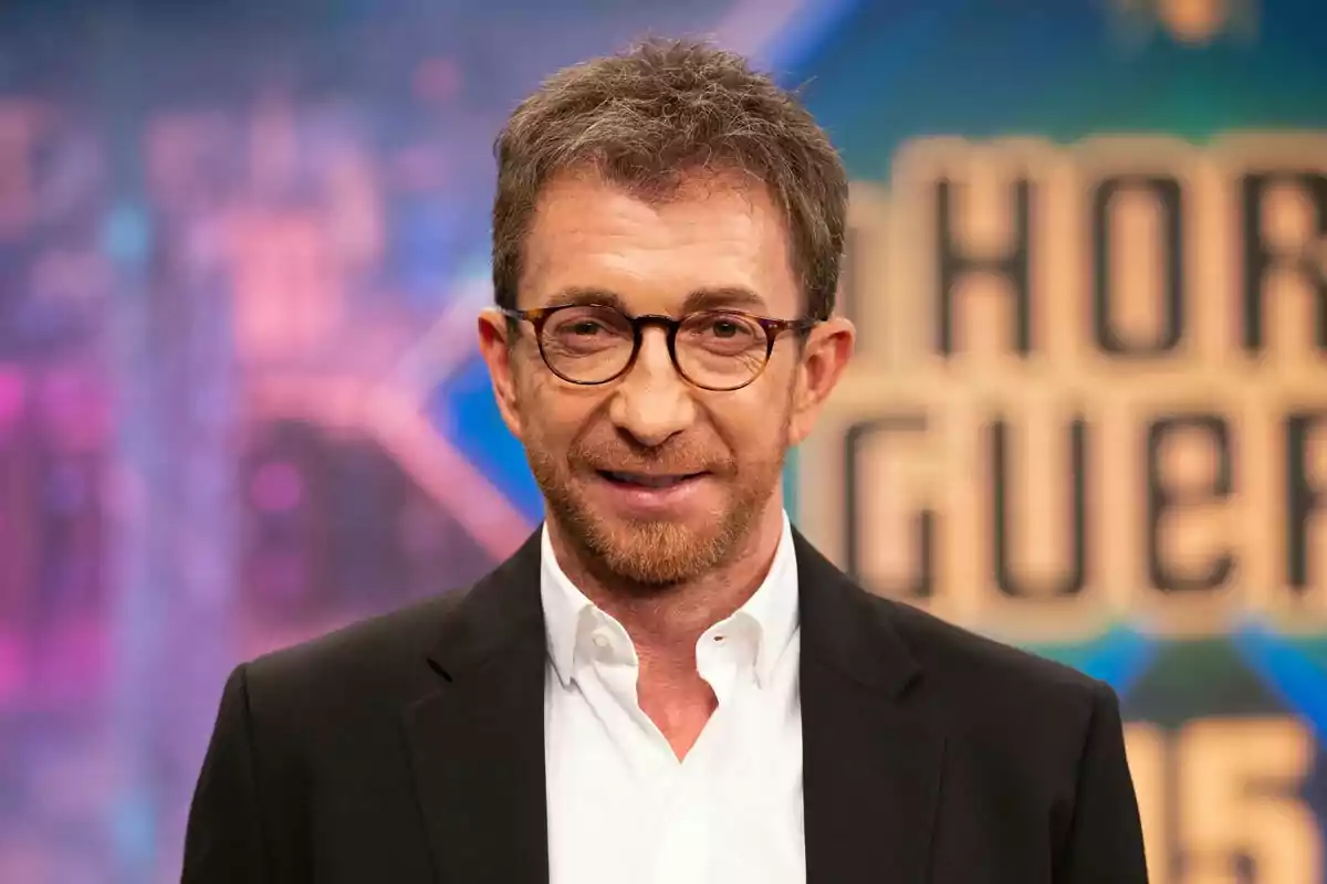 Hombre con gafas y barba corta vestido con camisa blanca y chaqueta oscura en un set de televisión colorido