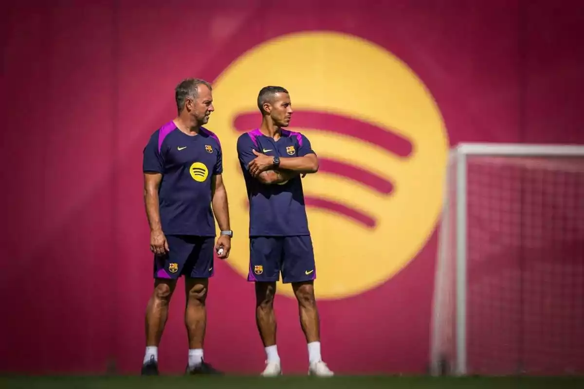 Two FC Barcelona coaches in training uniforms watch the field in front of a goal and a backdrop with the Spotify logo. Two FC Barcelona coaches in training uniforms watch the field in front of a goal and a backdrop with the Spotify logo.