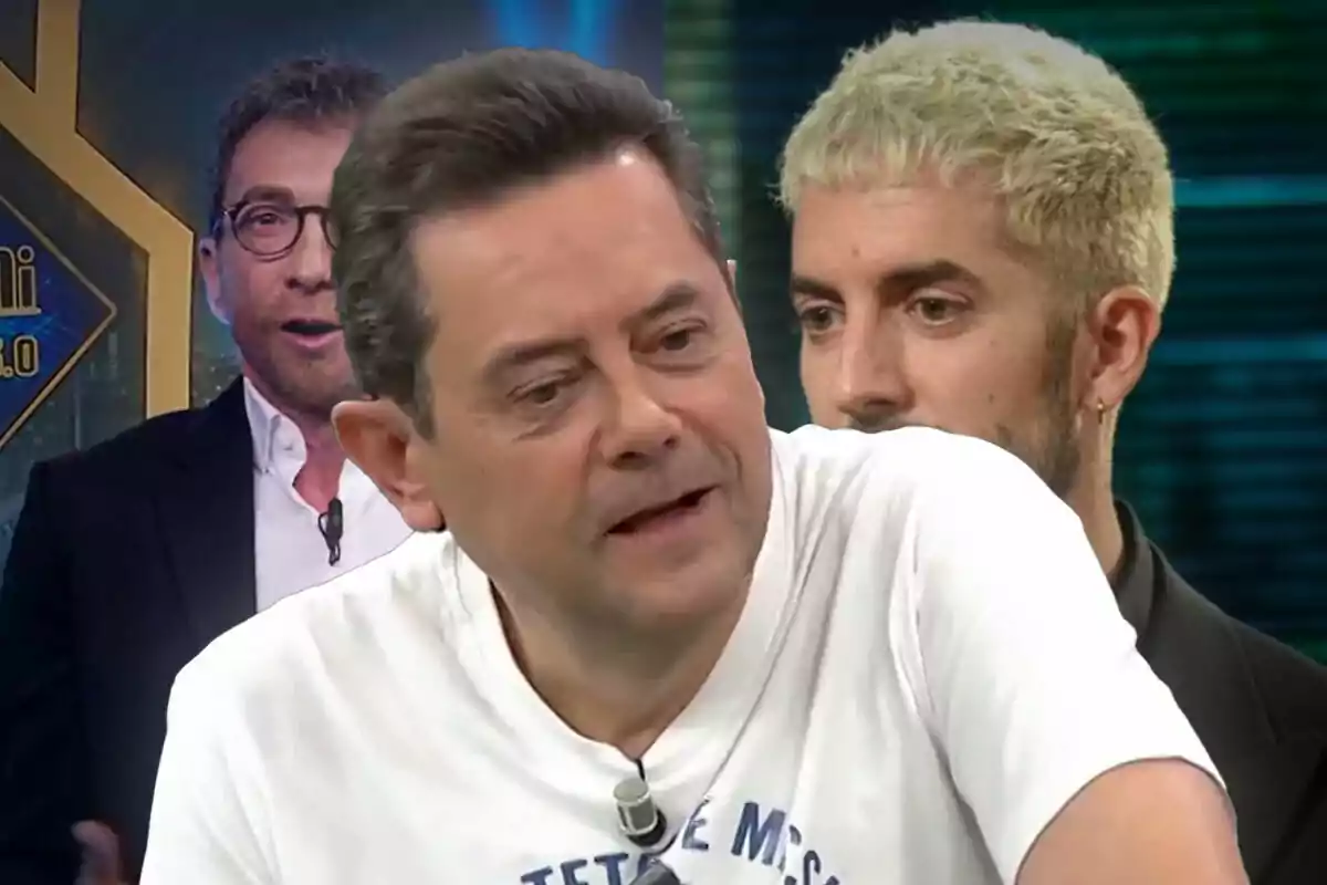 Tres hombres en un set de televisión, uno en primer plano con camiseta blanca, otro con cabello rubio corto y un tercero al fondo con gafas y chaqueta oscura