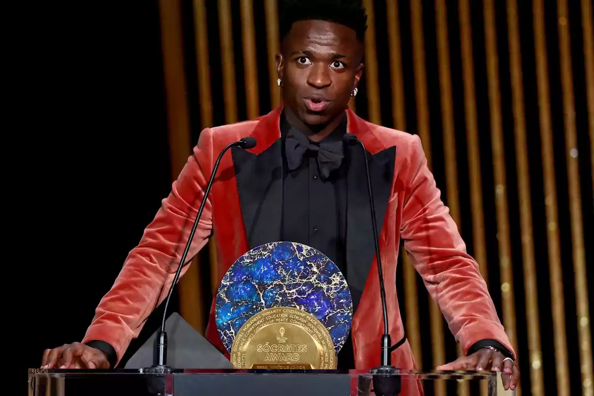 A person in a red and black tuxedo stands behind a podium with a decorative award in front of it.