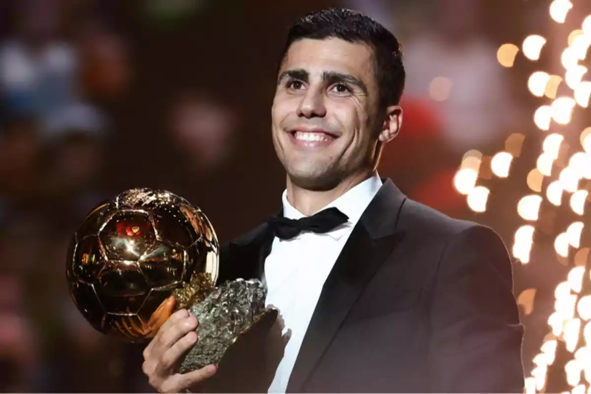 A smiling man in a suit holds a golden trophy with bright lights in the background.