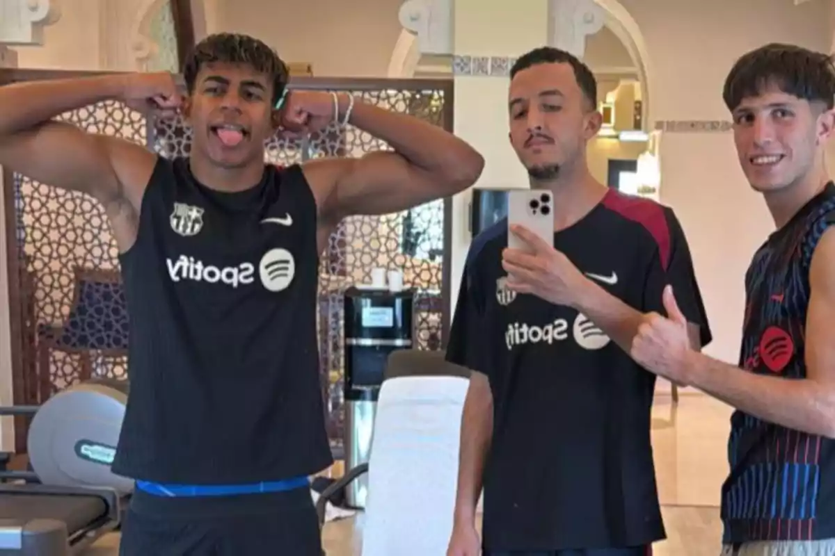 Three people pose in a gym, one of them flexing his arms and sticking out his tongue, another taking a selfie with a phone and the third giving a thumbs up, all wearing black sports jerseys with the Spotify logo.