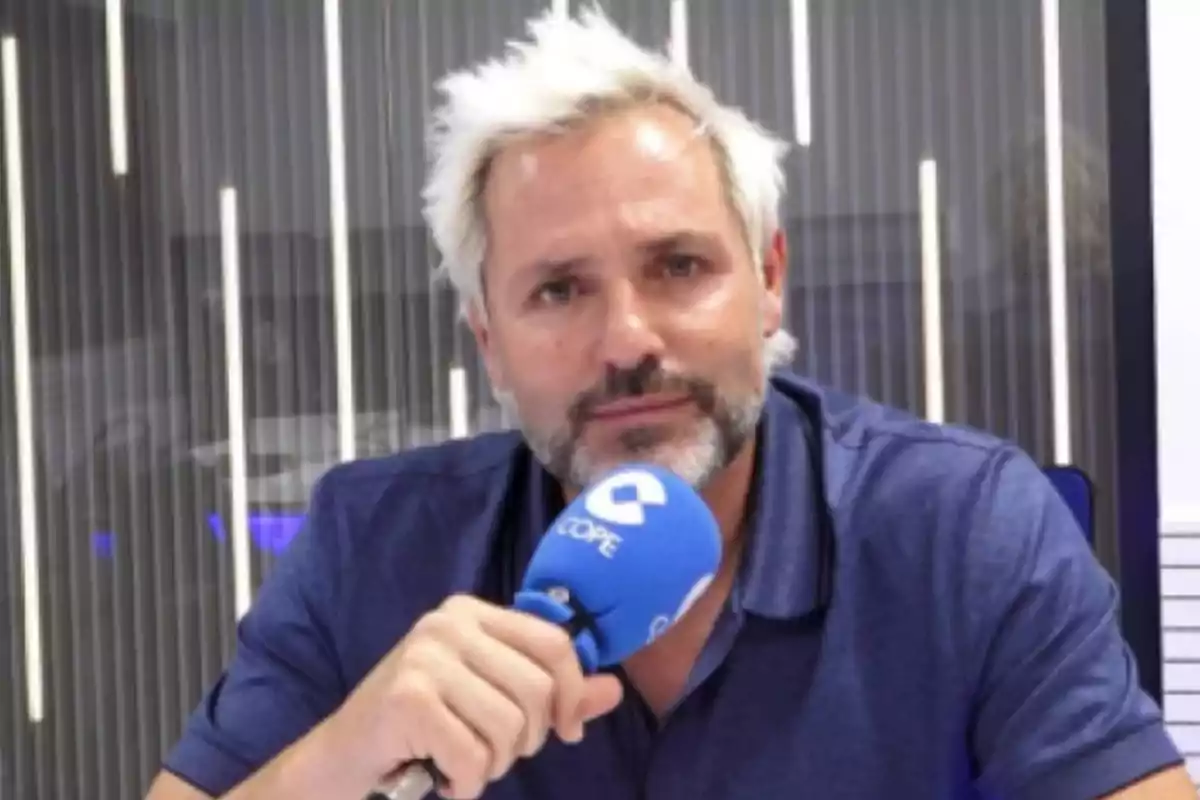 A man with light hair and a beard holds a blue microphone in a radio studio.