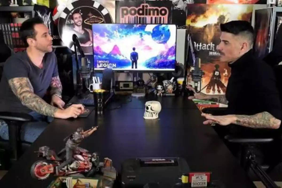 Two people with tattoos are talking in a studio with microphones and a screen displaying a colorful landscape, surrounded by decorative figures and posters.