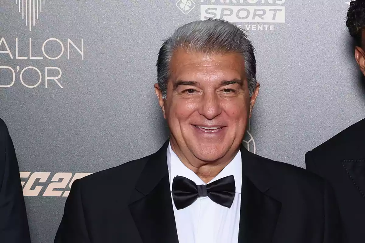 A man in a tuxedo and bow tie smiles in front of a background with logos. A man in a tuxedo and bow tie smiles in front of a background with logos.