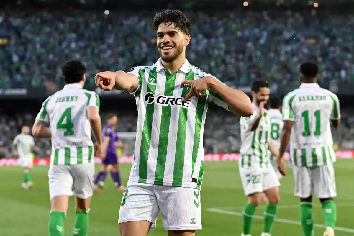Futbolista del Real Betis celebrando un gol en el campo junto a sus compañeros con la afición de fondo Futbolista del Real Betis celebrando un gol en el campo junto a sus compañeros con la afición de fondo