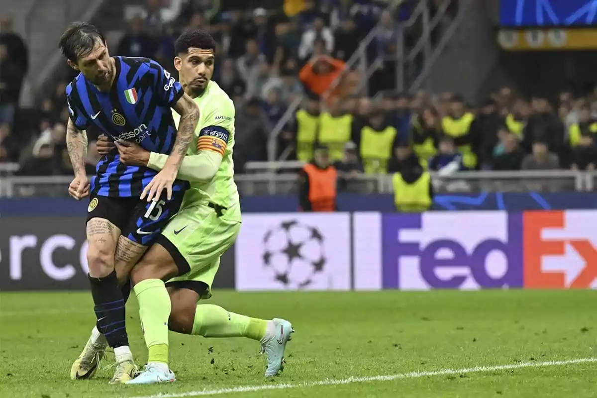 Dos jugadores de fútbol compiten por el balón en un partido, uno lleva uniforme azul y negro y el otro verde, con público y personal de seguridad en el fondo. Dos jugadores de fútbol compiten por el balón en un partido, uno lleva uniforme azul y negro y el otro verde, con público y personal de seguridad en el fondo.
