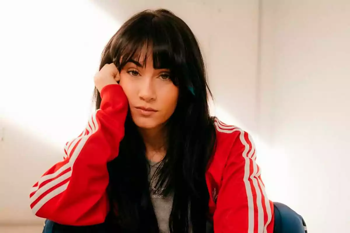 Aitana Ocaña with dark hair and bangs, wearing a red jacket with white stripes, resting her head on her hand. Aitana Ocaña with dark hair and bangs, wearing a red jacket with white stripes, resting her head on her hand.