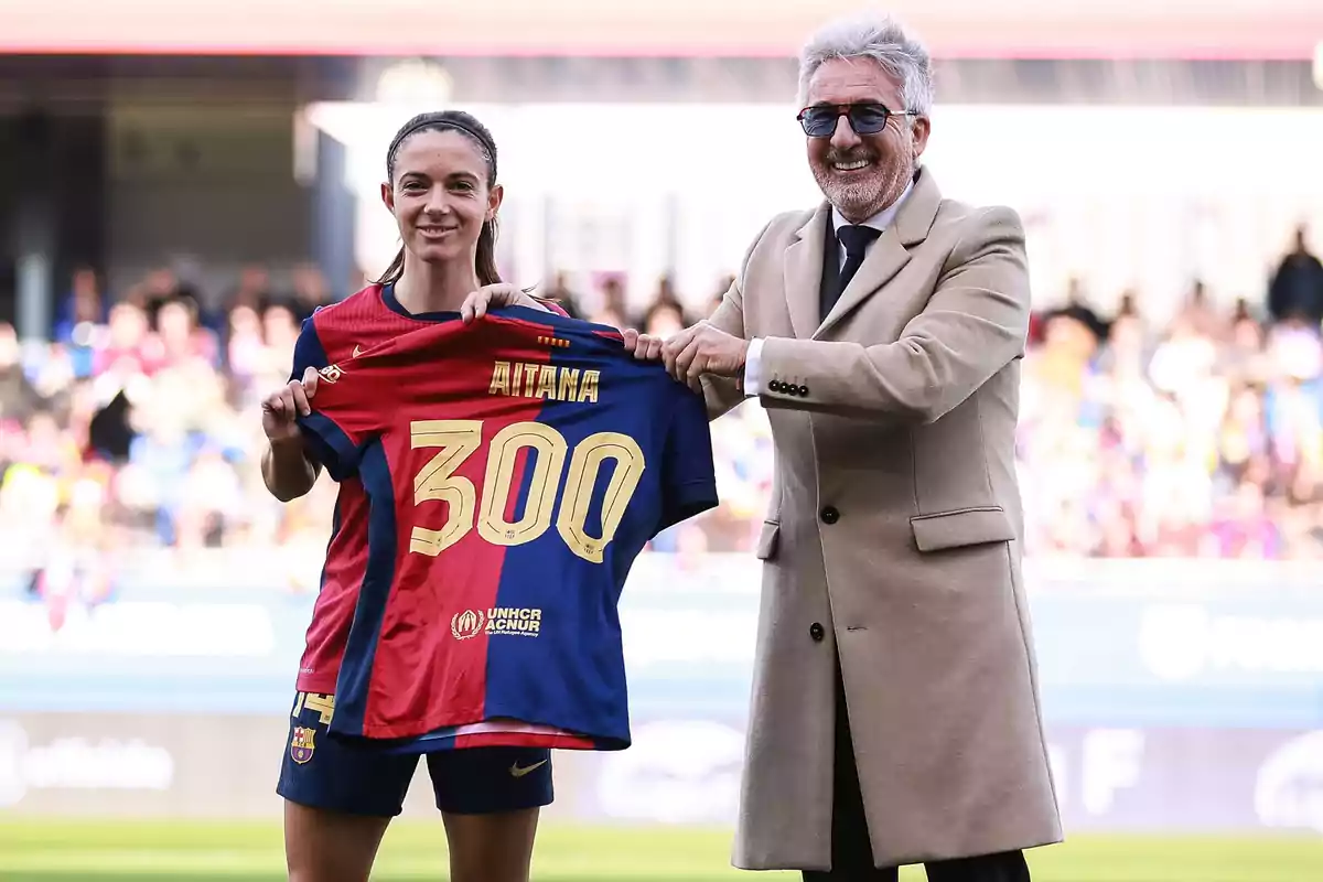 Una jugadora de fútbol y un hombre sonriente sostienen una camiseta con el número 300 en un estadio.