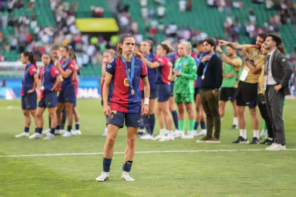 Soccer players in blue and red uniforms on the field, one of them in front with a medal and a serious expression, while the rest of the team and coaching staff are in the background.