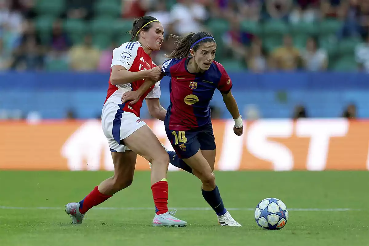 Dos jugadoras de fútbol compiten por el balón en un partido, una lleva el uniforme del Barcelona y la otra del Arsenal, en un estadio con público de fondo. Dos jugadoras de fútbol compiten por el balón en un partido, una lleva el uniforme del Barcelona y la otra del Arsenal, en un estadio con público de fondo.