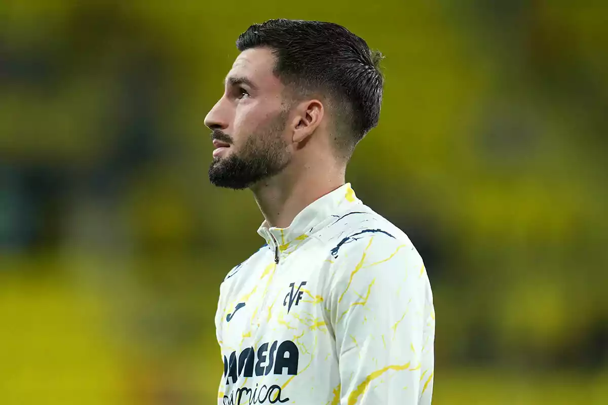 Álex Baena, con uniforme blanco y detalles amarillos, mira hacia arriba en un estadio con fondo desenfocado.