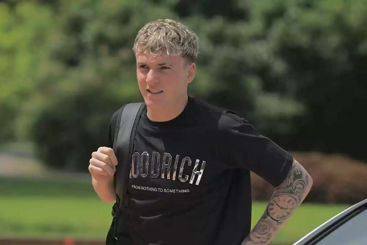 Álex Jiménez in a black t-jersey and backpack walks outdoors with trees in the background.