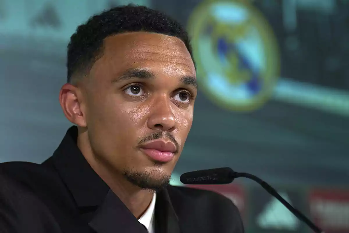Un hombre con traje oscuro habla en una conferencia de prensa con el escudo del Real Madrid desenfocado al fondo