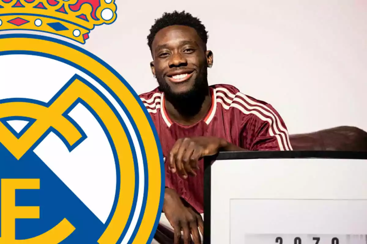 Un hombre sonriente con una camiseta deportiva roja y blanca junto a un gran escudo del Real Madrid.