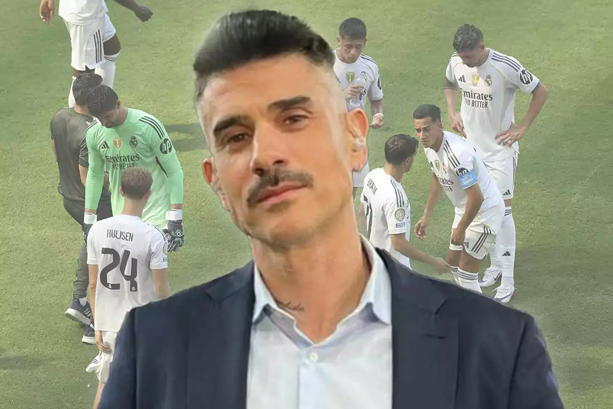 Un hombre de cabello corto y bigote aparece en primer plano con expresión seria mientras al fondo se observa a varios jugadores de fútbol vestidos con uniforme blanco en un campo de juego.