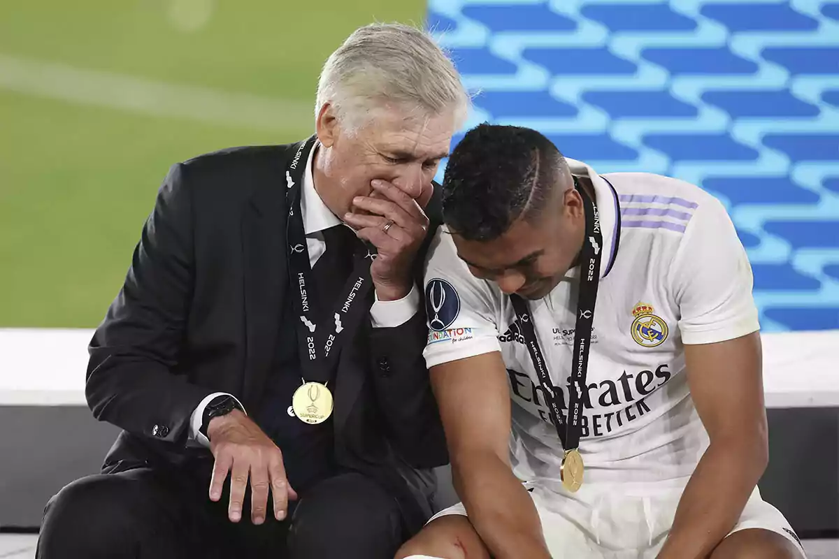 Two people sitting sharing a moment of complicity and laughter, both wearing medals and one of them wearing a suit while the other is wearing a sports uniform.