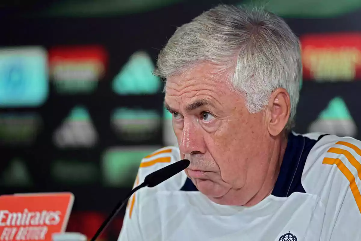 Carlo Ancelotti con expresión seria habla en una conferencia de prensa, con un micrófono frente a él y un fondo desenfocado con logotipos.