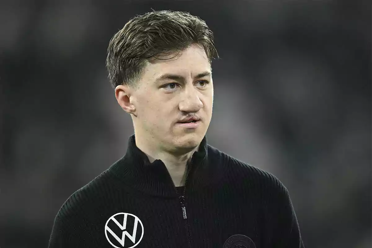 Un hombre con cabello corto y oscuro lleva un suéter negro con un logotipo circular en el pecho. Un hombre con cabello corto y oscuro lleva un suéter negro con un logotipo circular en el pecho.