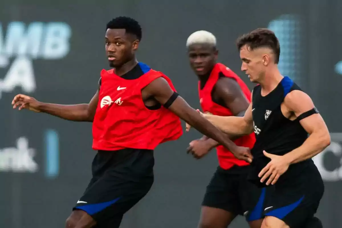 Soccer players training on the field, one of them is wearing a red bib while the other two are wearing black t-shirts.