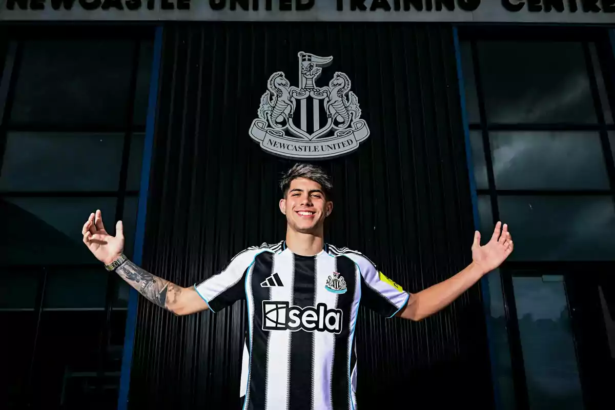 Player in Newcastle United uniform smiles and extends his arms in front of the club crest at the training center. Player in Newcastle United uniform smiles and extends his arms in front of the club crest at the training center.