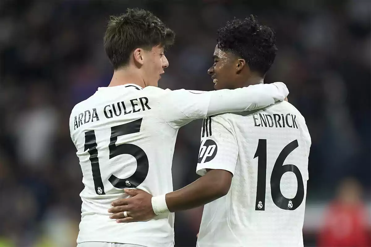 Dos jugadores de fútbol con camisetas blancas se abrazan en el campo.