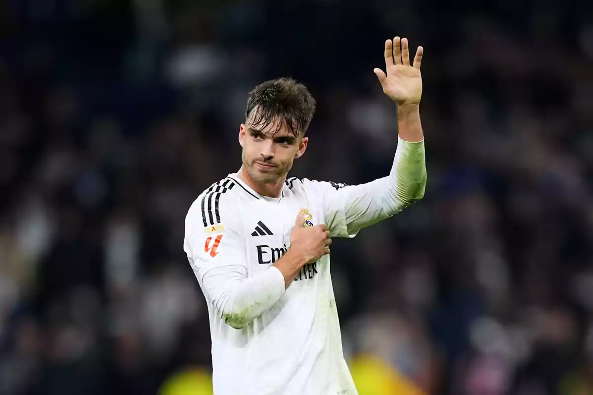 Un jugador de fútbol con uniforme blanco saluda levantando la mano en un estadio.