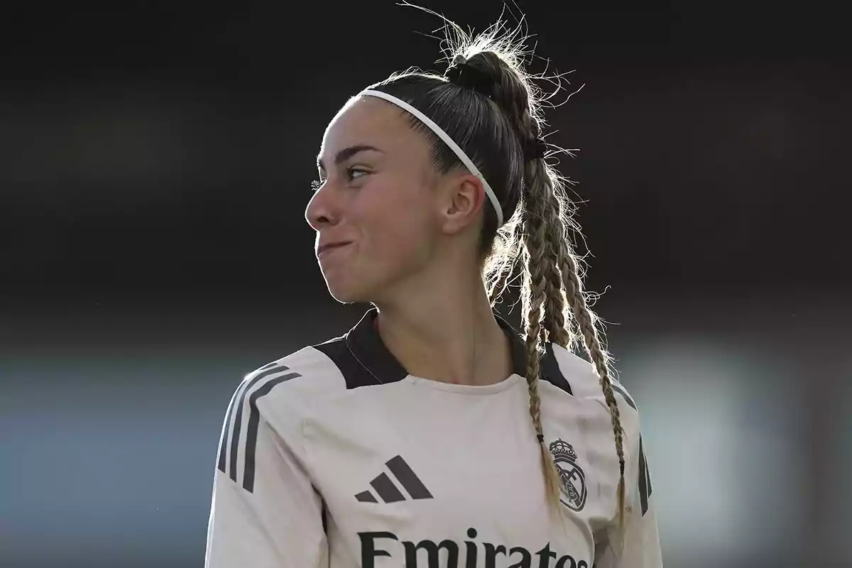 A female soccer player in a Real Madrid uniform looks to the side with a thoughtful expression.