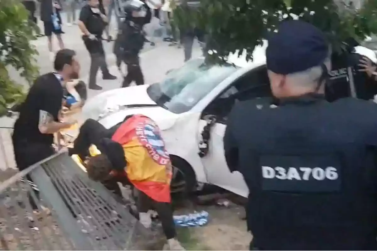 A group of people and police officers around a crashed white car on a street. A group of people and police officers around a crashed white car on a street.