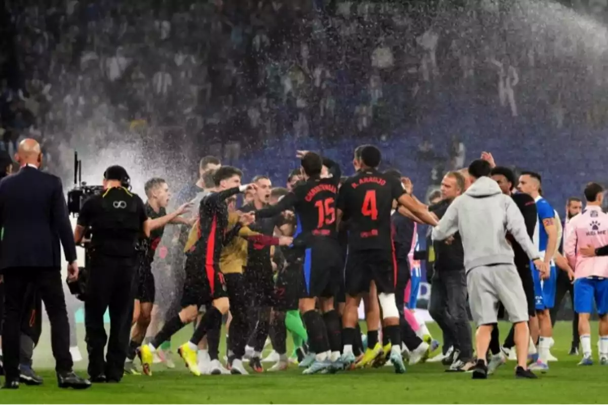 A group of players celebrates on the field while being sprayed with water, surrounded by cameras and technical staff. A group of players celebrates on the field while being sprayed with water, surrounded by cameras and technical staff.