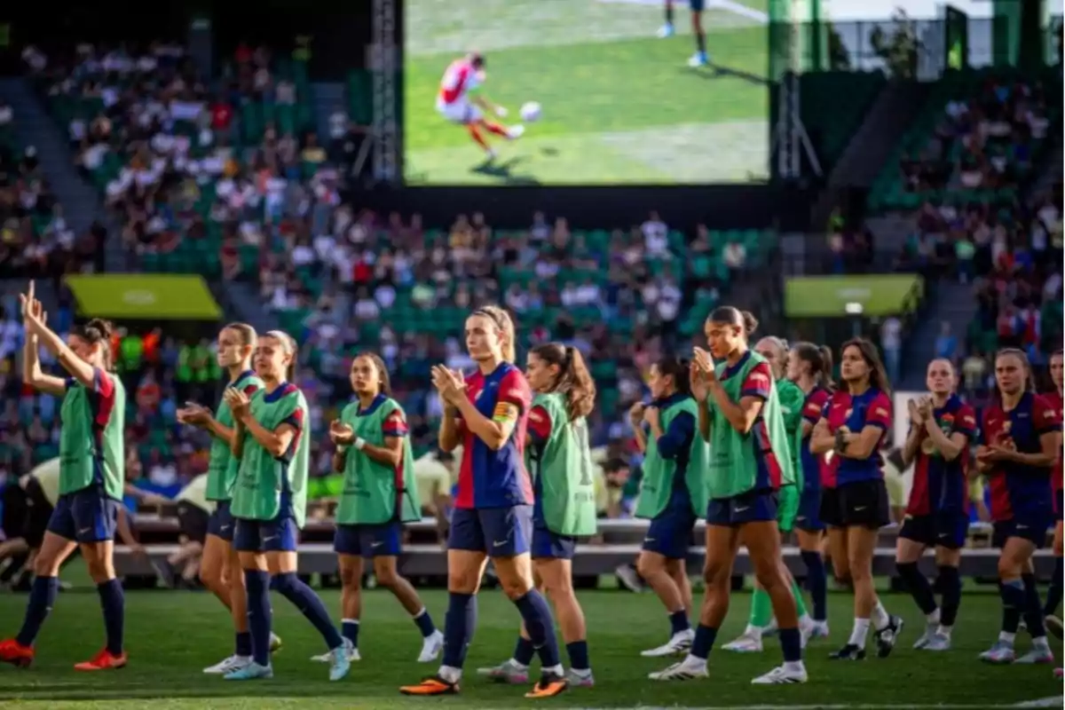 Jugadoras de fútbol aplauden en el campo mientras el público observa desde las gradas y una pantalla muestra una jugada. Jugadoras de fútbol aplauden en el campo mientras el público observa desde las gradas y una pantalla muestra una jugada.