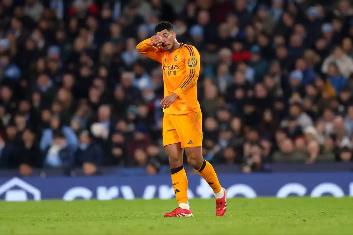 A player in an orange uniform wipes his face on the field during a match, with a crowd of spectators in the background.