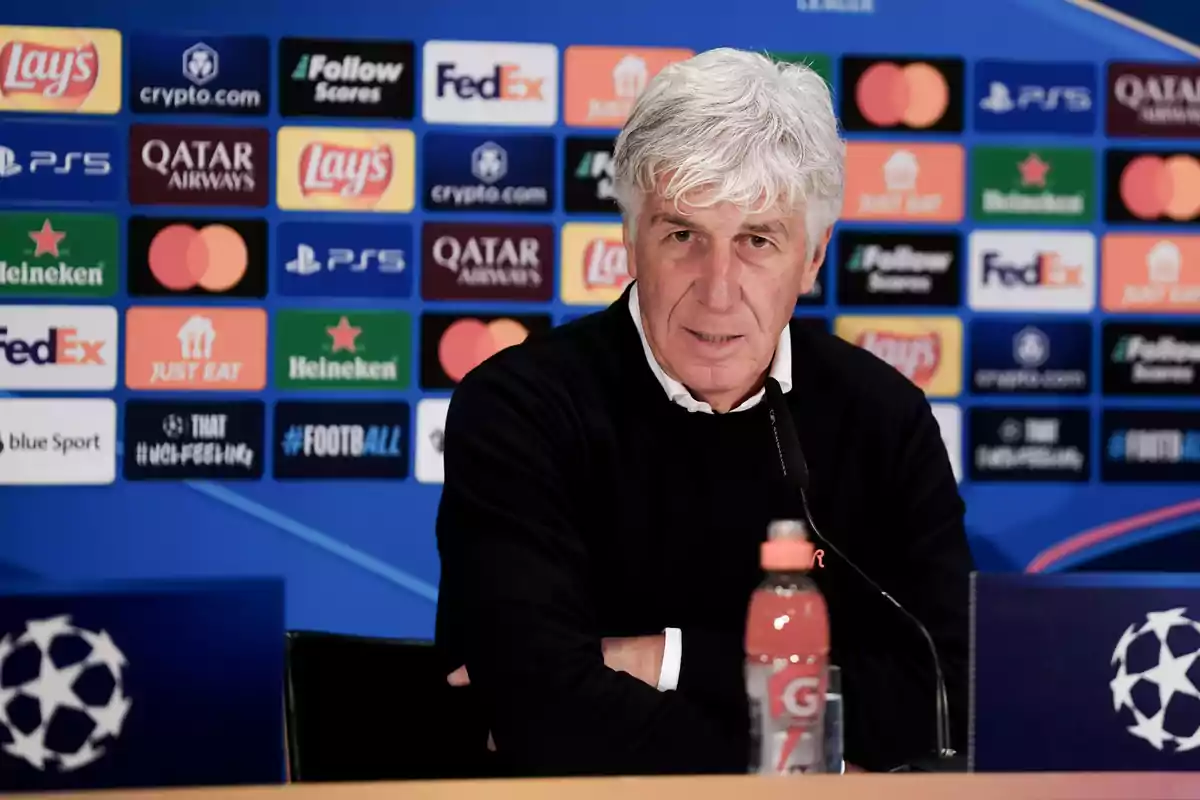 A gray-haired man at a press conference against a background of sports sponsor logos.