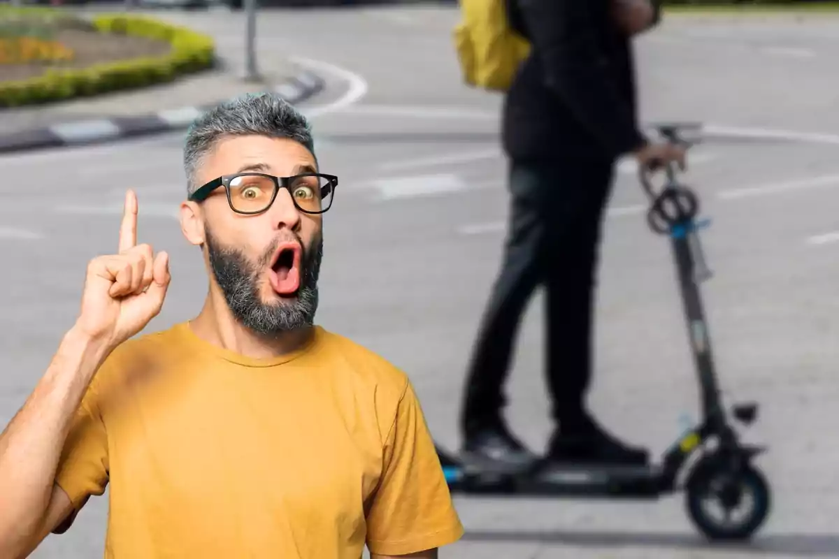 Un hombre con barba y gafas, vistiendo una camiseta amarilla, hace una expresión de sorpresa mientras señala hacia arriba con el dedo índice; al fondo, una persona en patinete eléctrico.