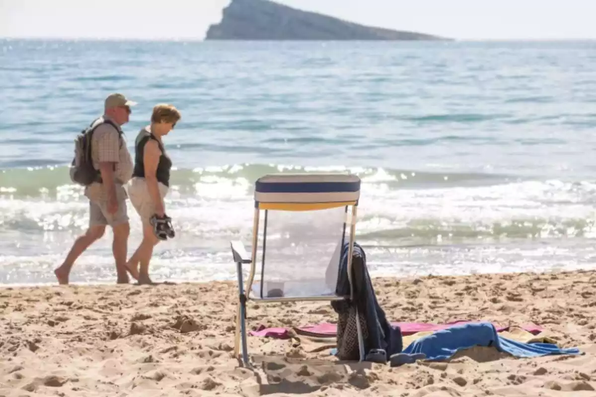 Dos personas mayores pasean a la orilla de la playa, con una silla vacía en la arena y el mar de fondo Dos personas mayores pasean a la orilla de la playa, con una silla vacía en la arena y el mar de fondo