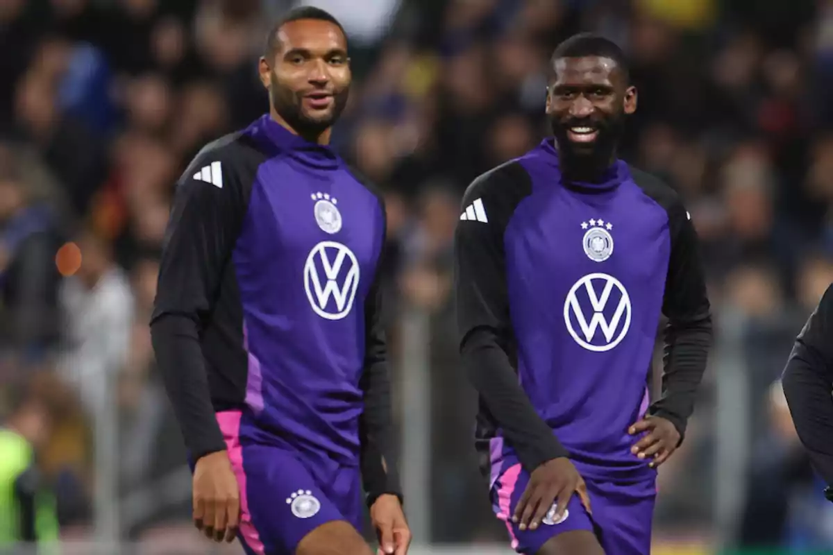 Two soccer players in purple and black training uniforms smile on the field.