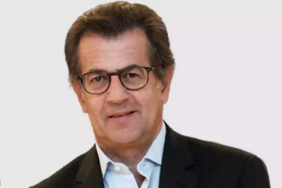A man with glasses and dark hair wears a dark suit and light jersey on a white background. A man with glasses and dark hair wears a dark suit and light jersey on a white background.