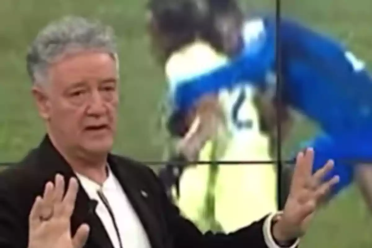 A man with gray hair and a serious expression is talking in front of a screen showing a blurred sports scene. A man with gray hair and a serious expression is talking in front of a screen showing a blurred sports scene.