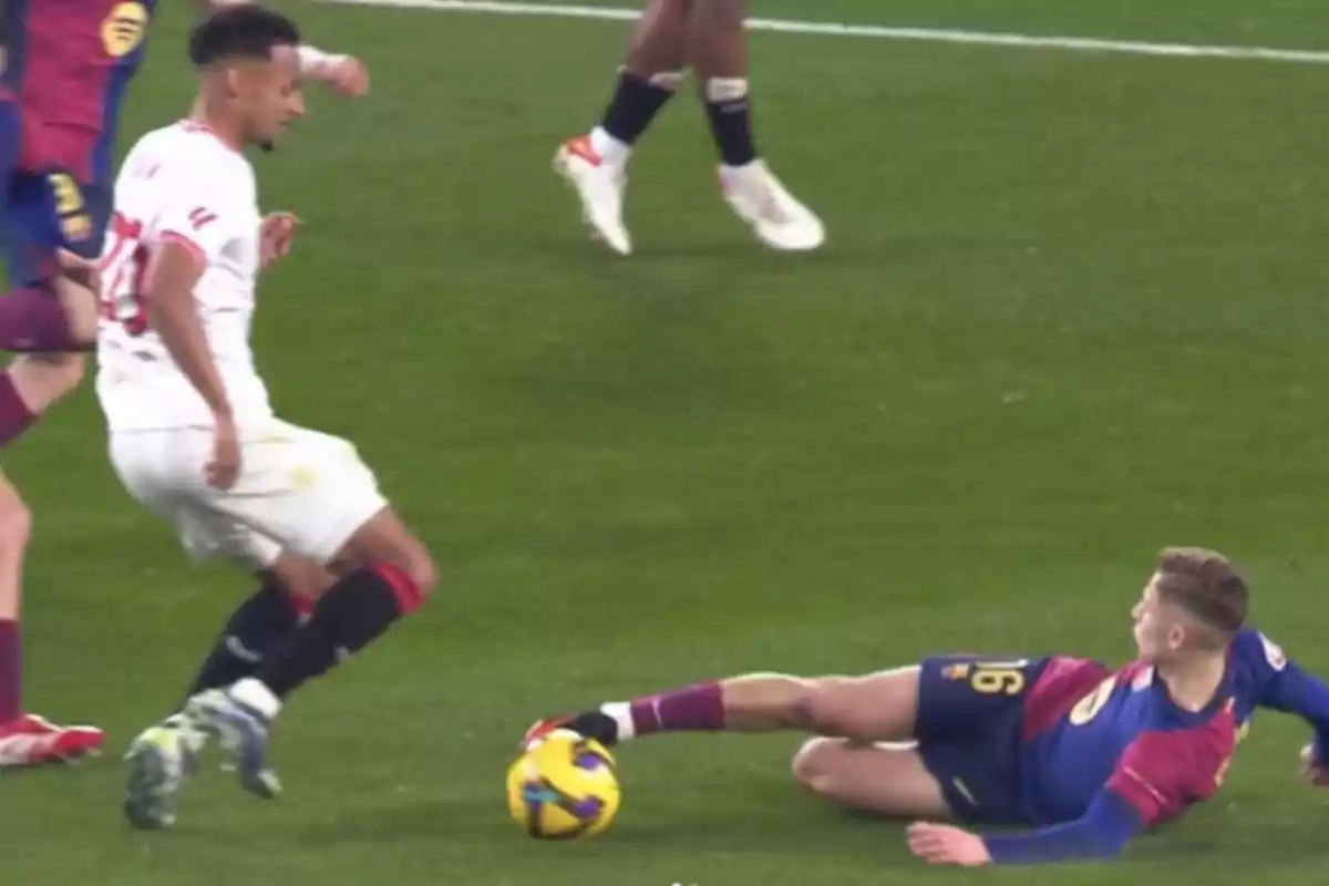 Two players in action during a match, one of them is on the ground trying to reach the ball with his foot while the other approaches with the ball at his feet.