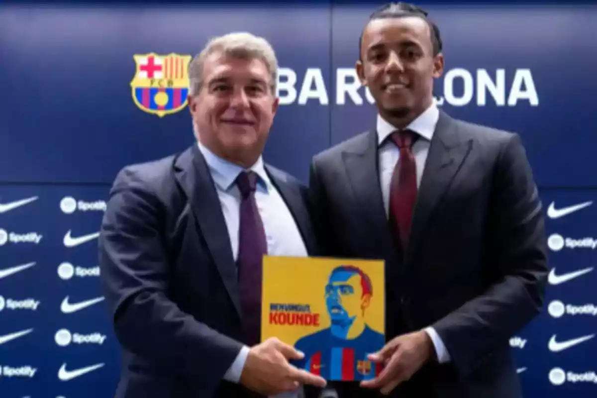 Two men in suits pose in front of an FC Barcelona backdrop, holding a colorful illustration.